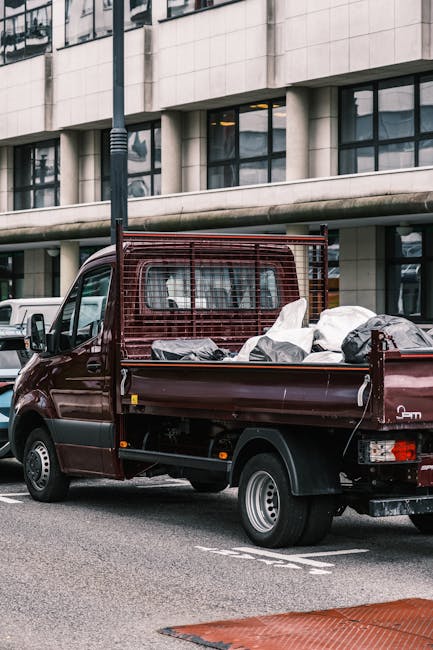 A burgundy flatbed van parked on a city street in front of a modern building with large glass windows and concrete panels. The vehicle's cargo area is filled with various household items including cardboard boxes, plastic-wrapped furniture, and large plastic bags, indicating a home relocation or furniture transport process. Some of the boxes and packages are stacked and secured for transit. The van is situated near a pavement with a streetlamp visible in the background, and other parked vehicles can be seen nearby. The scene appears to be during daylight hours, with natural light illuminating the area. This loading and packing activity aligns with professional removals and moving services provided by companies like Man with Van Hillingdon, specialising in efficient property relocations and waste removal, such as bulky items that need safe transportation and disposal.
