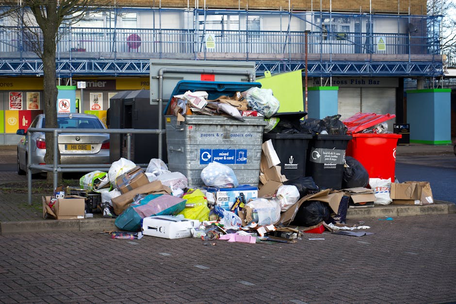 A collection of overflowing rubbish and recycling bins situated on a paved sidewalk outside a commercial area, with open cardboard boxes and plastic bags scattered around. The waste includes crumpled paper, cardboard packaging, plastic bottles, and miscellaneous household items, some spilling onto the street. The bins are positioned next to a metal railing, with a parked car and a building in the background. The scene appears to depict bulk waste collection, relevant to home or office relocation services offered by Man with Van Hillingdon, although the setting primarily focuses on waste removal rather than moving logistics.