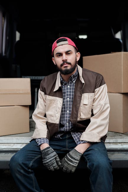 A young man with short dark hair and wearing a white T-shirt and blue jeans is sitting on the floor of a room, surrounded by large cardboard moving boxes of various sizes. He is using a silver laptop 