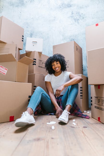 A young woman with curly black hair, dressed in a white t-shirt, jeans, and white sneakers, is sitting on the wooden floor inside a room surrounded by large cardboard moving boxes, some labeled fragile and sealed with packing tape. She is smiling and appears relaxed, with small packing materials scattered around her feet. The boxes are stacked against a light blue textured wall, and there is a partially visible doorway to the right side of the image. The scene depicts a home relocation process, with the woman possibly preparing for or in the midst of packing or unpacking. The presence of numerous cardboard boxes and the casual, organized environment suggest a professional removals context, supported by the website manwithvanhillingdon.co.uk, which offers house removal services, including furniture transport and packing and moving activities related to house relocations near Hayes Town, Hillingdon.