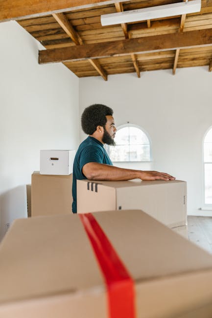 A man with a beard and short curly hair, wearing a blue t-shirt, stands inside a spacious room with a high wooden ceiling and exposed wooden beams. He is positioned behind several large cardboard boxes, some stacked on top of each other, with visible packing tape and a red parcel strap on one of the boxes. The room has white walls and two arched windows allowing natural light to illuminate the space. The man appears to be in the process of moving or organizing household items, consistent with home relocation or furniture transport activities. This setting reflects a typical home moving environment, with packaging materials and a focus on the loading or packing process, highlighting services offered by Man with Van Hillingdon related to removals and moving logistics near Hayes Town, Hillingdon.