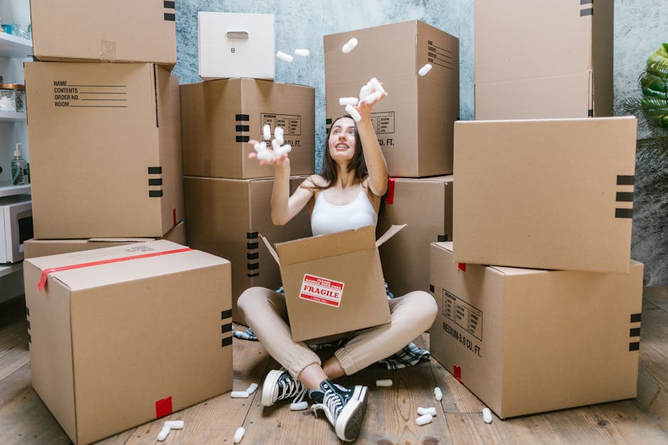 A young woman with curly black hair, dressed in a white t-shirt, jeans, and white sneakers, is sitting on the wooden floor inside a room surrounded by large cardboard moving boxes, some labeled fragile and sealed with packing tape. She is smiling and appears relaxed, with small packing materials scattered around her feet. The boxes are stacked against a light blue textured wall, and there is a partially visible doorway to the right side of the image. The scene depicts a home relocation process, with the woman possibly preparing for or in the midst of packing or unpacking. The presence of numerous cardboard boxes and the casual, organized environment suggest a professional removals context, supported by the website manwithvanhillingdon.co.uk, which offers house removal services, including furniture transport and packing and moving activities related to house relocations near Hayes Town, Hillingdon.