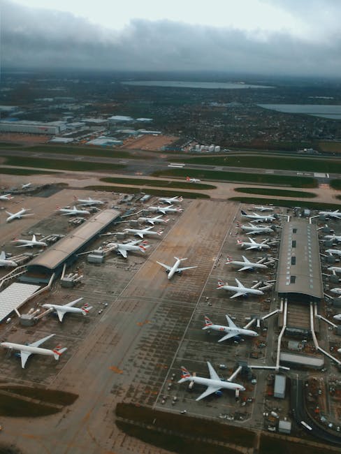 An aerial view of an airport tarmac showing numerous commercial airplanes parked along the apron, with some positioned near gates connected to terminal buildings. The aircraft are mainly white with red and dark blue branding on their tails. The tarmac is marked with yellow and white lines, and areas of concrete and asphalt are visible. In the background, there are runways and a landscape of fields, industrial areas, and water bodies under a cloudy sky. This scene emphasizes airport infrastructure and its connection to logistics, relevant to home relocation and furniture transport services provided by Man with Van Hillingdon, especially in relation to Heathrow-area moves involving airport access and parking considerations.