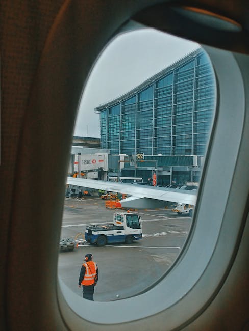 View through an airplane window showing the airport tarmac with ground crew personnel in high-visibility orange vests and dark clothing, operating a small tug vehicle near a parked airplane. The airplane is connected to a jet bridge leading to a modern terminal building with a glass facade, which has a large number 522 displayed near the entrance. The scene involves aircraft loading and ground support activities, with clear weather and diffused daylight. This image illustrates the logistics and movements involved in home relocation and furniture transport services offered by Man with Van Hillingdon at Heathrow airport, highlighting the importance of efficient packing, loading, and airport access during house removals.