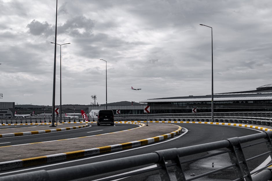 An aerial view of an airport tarmac showing numerous commercial airplanes parked along the apron, with some positioned near gates connected to terminal buildings. The aircraft are mainly white with red and dark blue branding on their tails. The tarmac is marked with yellow and white lines, and areas of concrete and asphalt are visible. In the background, there are runways and a landscape of fields, industrial areas, and water bodies under a cloudy sky. This scene emphasizes airport infrastructure and its connection to logistics, relevant to home relocation and furniture transport services provided by Man with Van Hillingdon, especially in relation to Heathrow-area moves involving airport access and parking considerations.