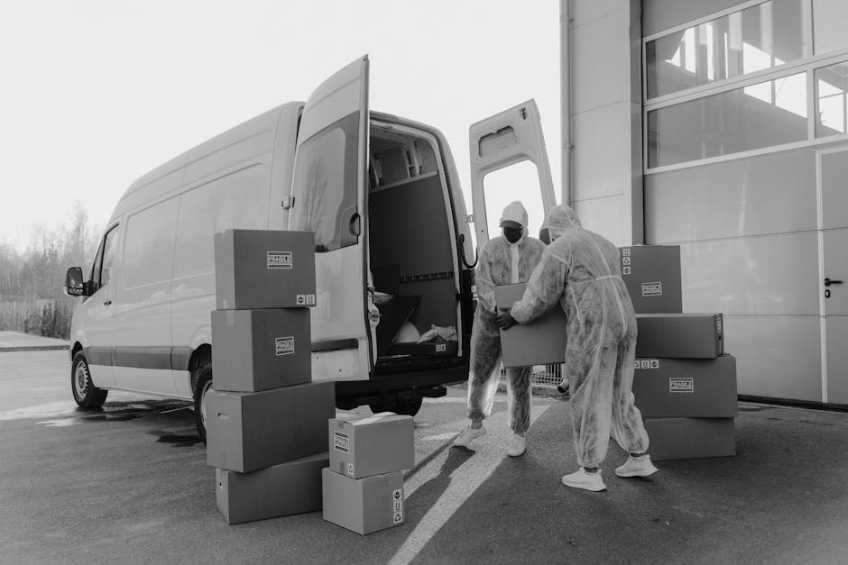 Two movers dressed in protective clothing, including white coveralls, face masks, and hair caps, are loading large cardboard boxes onto a white moving van parked outside a modern building with glass panels. The boxes vary in size and are stacked on the pavement and nearby, with some being carried by the movers and others already positioned near the van’s open rear door. The scene takes place during daylight, with natural light illuminating the area, indicating a home relocation or packing and moving process. The moving van, equipped for furniture transport and packing logistics, is positioned adjacent to the building entrance, with additional boxes stacked on the ground around it. Man with Van Hillingdon, a professional removals service, is involved in a house moving operation, supporting efficient loading during the home relocation process, consistent with Hillingdon Council moving van permit rules.