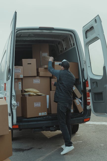 Two movers dressed in protective clothing, including white coveralls, face masks, and hair caps, are loading large cardboard boxes onto a white moving van parked outside a modern building with glass panels. The boxes vary in size and are stacked on the pavement and nearby, with some being carried by the movers and others already positioned near the van’s open rear door. The scene takes place during daylight, with natural light illuminating the area, indicating a home relocation or packing and moving process. The moving van, equipped for furniture transport and packing logistics, is positioned adjacent to the building entrance, with additional boxes stacked on the ground around it. Man with Van Hillingdon, a professional removals service, is involved in a house moving operation, supporting efficient loading during the home relocation process, consistent with Hillingdon Council moving van permit rules.