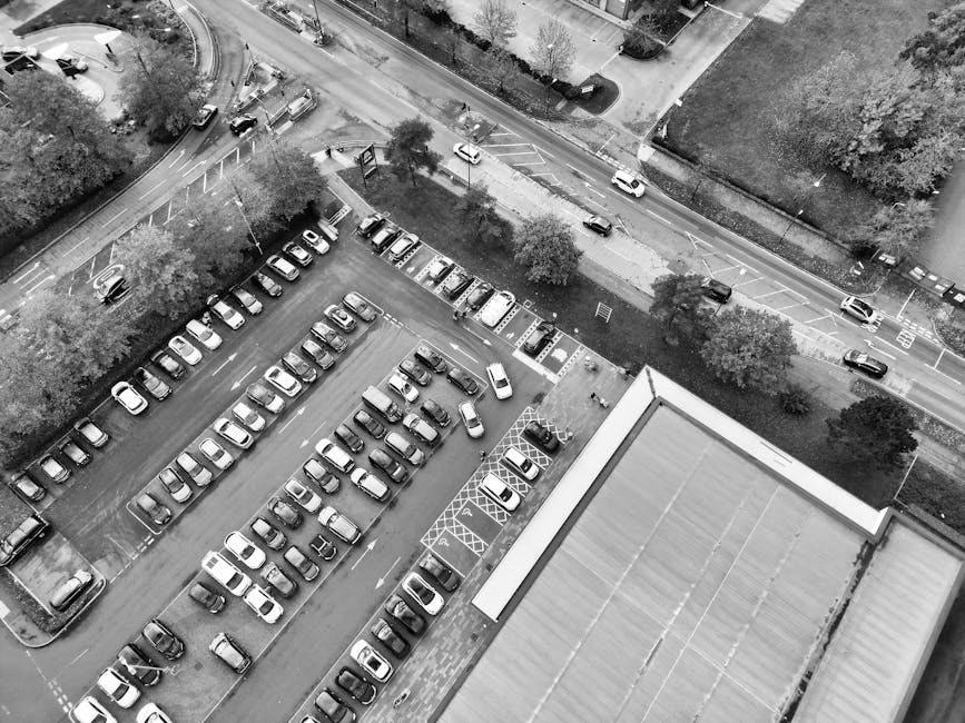 An aerial black and white photograph showing a large parking lot filled with numerous parked cars arranged in neat rows, with some empty spaces visible. To the left, a section of the parking lot is bordered by trees and a small green area. Adjacent to the parking lot is a road with several moving vehicles and a sidewalk lined with trees. Visible in the bottom right corner is the upper roof of a building, likely part of a commercial or residential property. Near the building, a trolley or dolly is seen carrying a few cardboard boxes and large wrapped furniture items, indicating a home removal or furniture transport process. The scene suggests a location with scheduled parking suspensions for moving services, possibly managed by Man with Van Hillingdon, as part of their house relocation logistics through their removals service category. The lighting indicates daytime conditions, with shadows cast by vehicles and trees, illustrating a typical urban environment prepared for a moving operation.