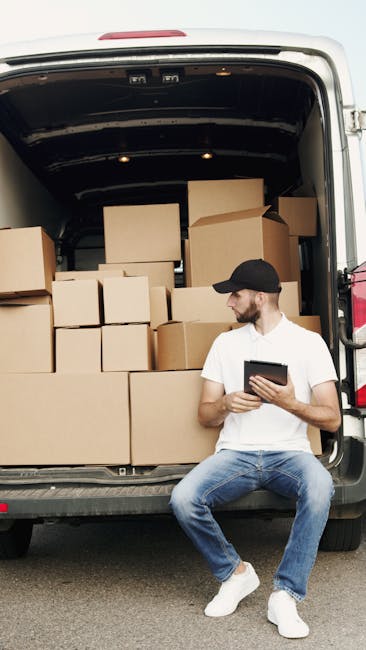 A young woman with curly hair is sitting at a table inside a bright room, closely examining a laptop placed on a large cardboard box, which is part of a home relocation packing process. Behind her, a