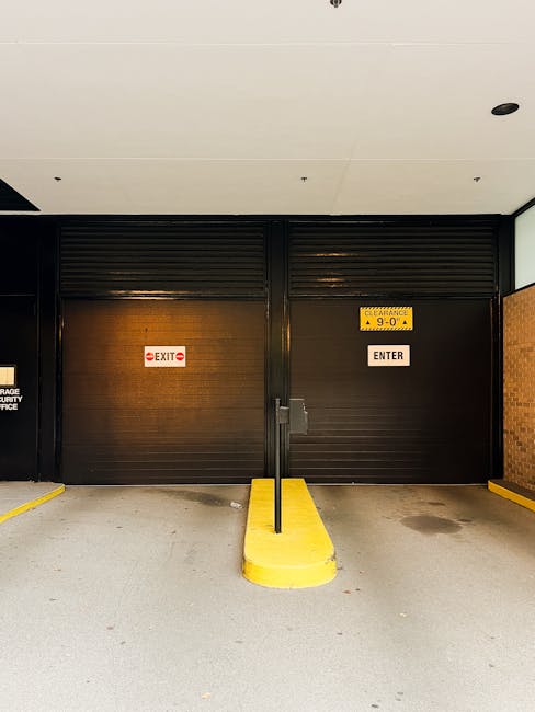 A black metal roller shutter door in an indoor parking/loading area with an 'EXIT' sign on the left side and an 'ENTER' sign on the right side. The right side of the door features a yellow sign indica