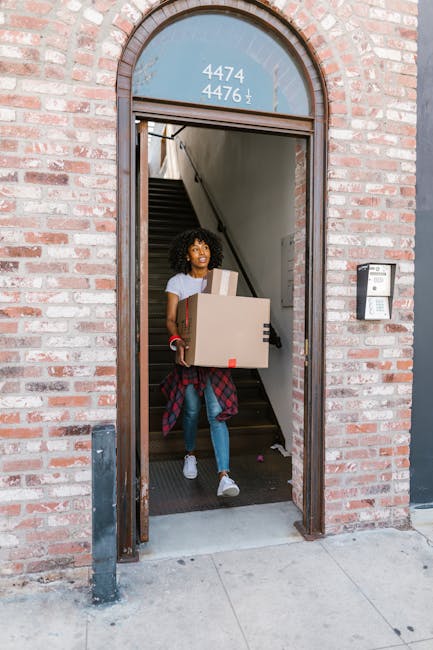A young woman with curly hair, wearing a light grey t-shirt, blue jeans, and white sneakers, is exiting a residential building through an open brick doorway. She is carrying a large cardboard box in her arms, bundled with protective packing materials, possibly bubble wrap or plastic wrap. Inside, visible stairs lead upward, and she appears to be engaged in a house removal or furniture transport process. The doorframe is made of dark metal, with a curved top section displaying the building's street number and address, and a wall-mounted intercom system is positioned on the right side of the entrance. The scene is set during daylight hours, with natural light illuminating the exterior pavement and doorway, reflecting typical moving or packing activities associated with home relocations. Occasionally, Man with Van Hillingdon offers professional removals services, often involving packing, loading, and transporting household items efficiently through areas like Ruislip HA4, where tight staircases may require careful handling during furniture transport.