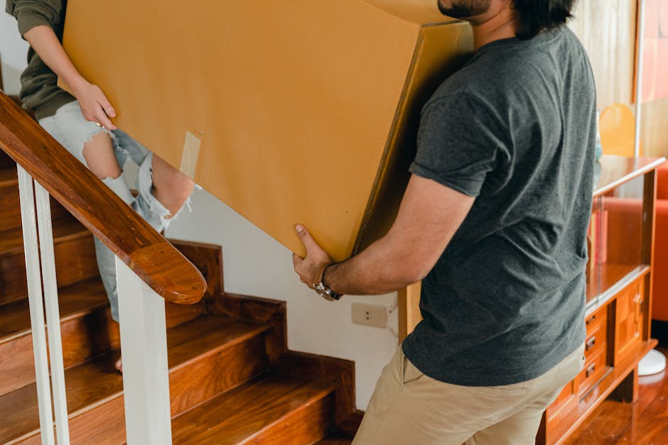 A man wearing a dark grey t-shirt and beige trousers is lifting a large, rectangular cardboard box with reinforced edges, likely containing household items or furniture, during a home relocation. He is positioned on a wooden staircase with polished dark wood steps and white-painted risers, holding the box securely with both hands. Inside the property, natural light illuminates the scene, with a glimpse of a partly visible person on the stairs wearing ripped jeans, assisting in the move. Nearby, a wooden sideboard with shelves and decorative items is visible against the wall, and a doorway with a curving stair bannister made of matching polished wood is nearby. The image captures the careful and coordinated process of furniture transport and packing during a moving operation, with a focus on handling large items carefully to navigate tight spaces, typical of a professional house removal service. Man with Van Hillingdon provides these relocation services, emphasizing careful loading and transport, especially for flats with limited stairway clearance, as discussed in the 'Ruislip HA4 flats: moving tips for tight staircases' guide.