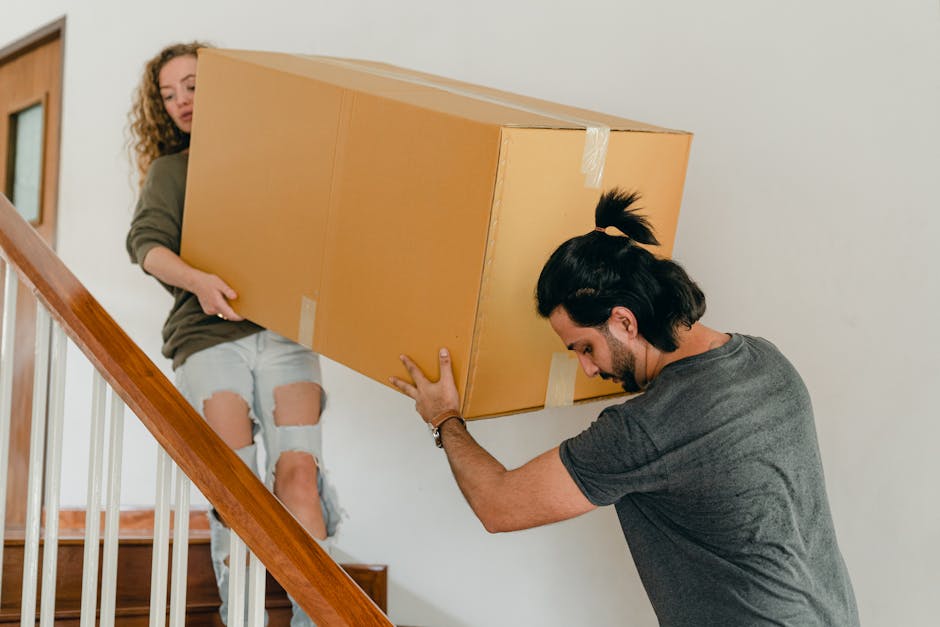 A young woman with curly hair, wearing a light grey t-shirt, blue jeans, and white sneakers, is exiting a residential building through an open brick doorway. She is carrying a large cardboard box in her arms, bundled with protective packing materials, possibly bubble wrap or plastic wrap. Inside, visible stairs lead upward, and she appears to be engaged in a house removal or furniture transport process. The doorframe is made of dark metal, with a curved top section displaying the building's street number and address, and a wall-mounted intercom system is positioned on the right side of the entrance. The scene is set during daylight hours, with natural light illuminating the exterior pavement and doorway, reflecting typical moving or packing activities associated with home relocations. Occasionally, Man with Van Hillingdon offers professional removals services, often involving packing, loading, and transporting household items efficiently through areas like Ruislip HA4, where tight staircases may require careful handling during furniture transport.