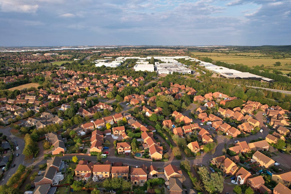 An aerial view of a residential neighbourhood showing rows of single-family houses with pitched roofs, surrounded by green trees and winding streets. To the background, there are large industrial warehouses with flat roofs, situated on open fields under a partly cloudy sky. The image captures a typical home relocation scene as part of an illustration for furniture transport and moving logistics, with the houses neatly arranged along curved roads and some driveways having parked cars. The lighting indicates daytime, providing clear visibility of the different building materials, including brick and tile roofs, as well as the landscaped gardens. This context relates to professional removals services, such as those offered by Man with Van Hillingdon, supporting local house moves and packing processes within suburban and industrial areas.